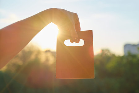 Paper bag for shopping in womans hand, bag made of brown recycled craft paper, background sunset, evening cityの写真素材