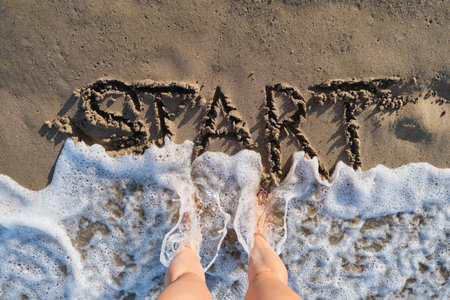 Text start written in sand on sea beach and womans bare feet. Sea foamy wave washing off the word...