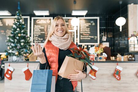 Happy smiling blonde in coffee shop with shopping bags and red poinsettia christmas flower. Winter portrait, girl in woolen scarf, with Christmas shopping.の写真素材