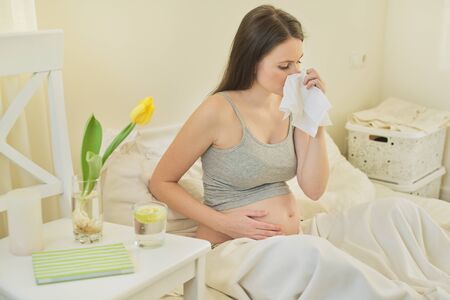 Young pregnant woman with handkerchief sitting in bed at home, sneezing and coughing, allergy cold seasonの写真素材