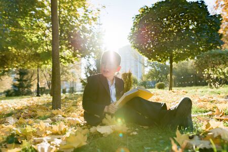 Outdoor autumn portrait of schoolboy reading book, background of yellow trees in the park, boy in jacket, golden hour, back to schoolの写真素材