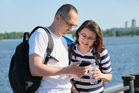 Outdoor mature couple using smartphone, man and woman talking walking in the park, people in...