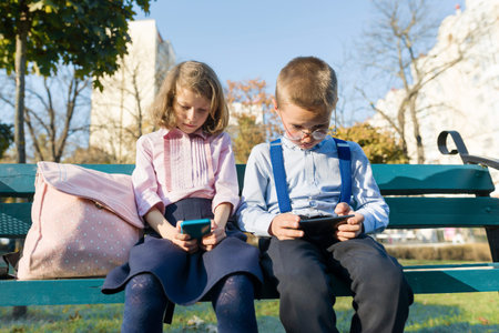 Smart serious children boy and girl are looking into smartphones. On a bench with school backpacks,...
