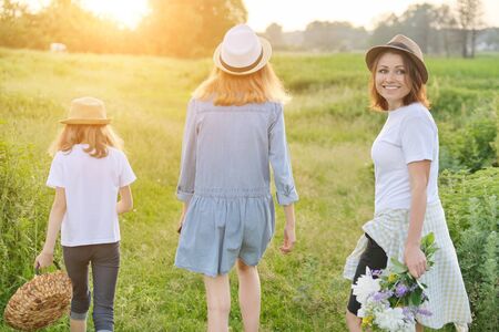Mother with children two daughters walking along a country road, background summer meadow sunset, back viewの写真素材
