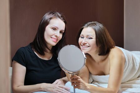 Talking and smiling women beautician and mature female patient with mirror in beauty salon. People, spa, cosmetology and skincare conceptの写真素材