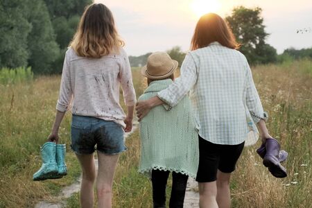 Family mother and two daughters walking together along country road after summer rain holding rubber boots in hands, back view, nature background golden hourの写真素材