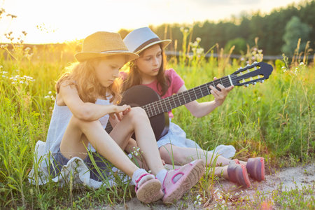 Children sitting in nature with classical guitar, two girls learning playing the guitar and singing, enjoying music and summer dayの写真素材