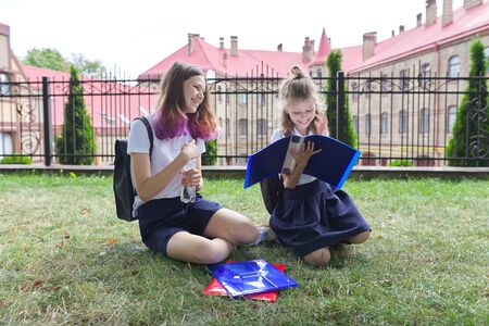 Two schoolgirls sitting on grass with books near school building, girls sisters, teenager and elementary school student laugh and look at textbooksの写真素材