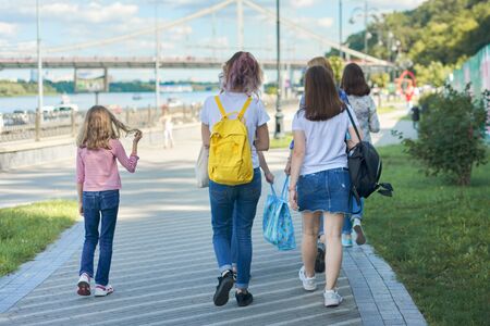 People walking, back view, teenage girls and child walk along sidewalk near highway and city promenadeの写真素材