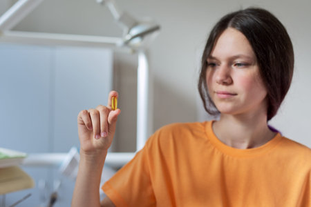 Teenage girl holding yellow vitamin d gel capsule in hand, dentist office background. Nutritional...