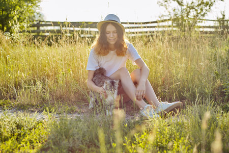 Teenager girl in hat on nature playing with gray fluffy cat, countryside, girl and pet on rural landscape, sunny summer dayの写真素材