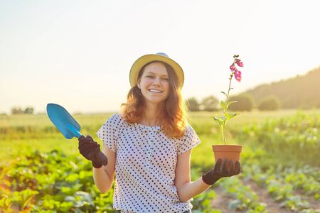 Young teenager girl in gloves walking with garden trowel and pot flower. Sunny spring summer day, rustic style, beautiful landscape backgroundの写真素材
