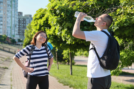 Mature smiling man and woman in sportswear with backpacks exercise mat walking in city park talking...