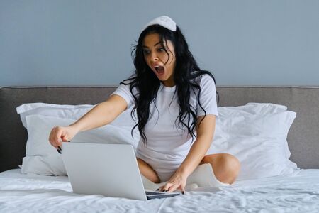 Beautiful emotional woman with open mouth sitting in bed using laptop computer, reaction of surprise on the face of femaleの写真素材