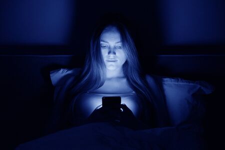 Young beautiful woman at night sitting in bed with smartphone. Smiling girl reading phone in dark. Lifestyle and technology in lives of young peopleの写真素材