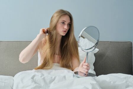 Young beautiful girl combing long hair with brush and mirror. Female sitting in bed, hair beauty care healthの写真素材