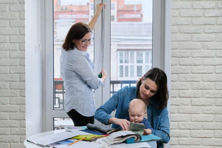 Window decoration with curtains, woman designer taking window sizes. At the table, sitting mother and baby choosing fabrics, color, modelの写真素材