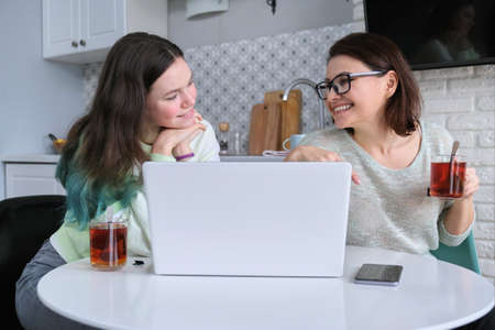 Communication mother and daughter teenager, talking parent and child sitting at home in kitchen and looking at laptop screenの写真素材
