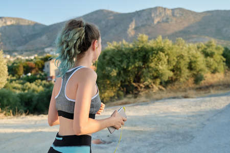 Running fitness girl in headphones with smartphone, back view, copy space. Sunny summer day, road in the mountains, active healthy lifestyle in young peopleの写真素材