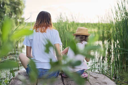 Children resting near the water on sunny summer day, two girls looking at lake sitting on bridge, relaxing, back viewの写真素材