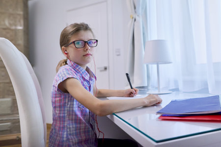 Girl child 9, 10 years old sitting at home at desk near window and writing in notebook, headphones...