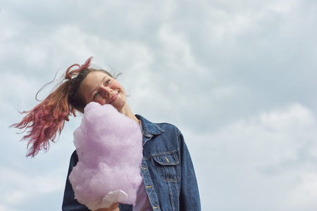 Beautiful smiling teenage girl with pink hair, hairstyle holding candy floss. Blue sky background,...