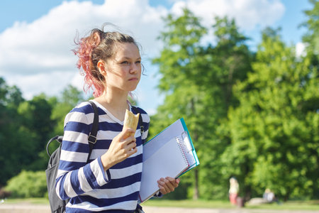 Girl student walking outdoor with backpack, notebooks, eating sandwich.
