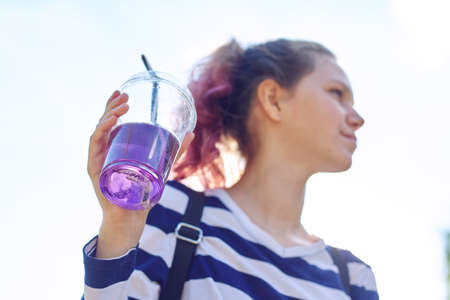 Teen girl holding glass with straw with purple drink in hand. Female quenching thirst on sunny summer day, sky background, copy spaceの写真素材