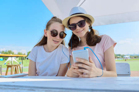 Two teenage girls in headphones listening music on smartphone, having fun on sunny summer day. Youth, teens, friendship, communication, people conceptの写真素材