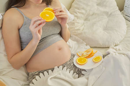 Young pregnant woman eating oranges sitting at home in bed. Natural vitamins, healthy foods, nutrition for pregnant womenの写真素材
