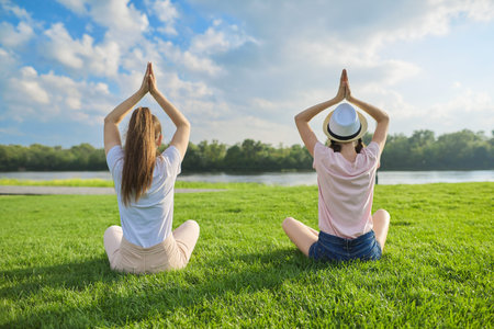 Two girls sitting in lotus position on green grass, back view, meditation on beautiful landscape,...
