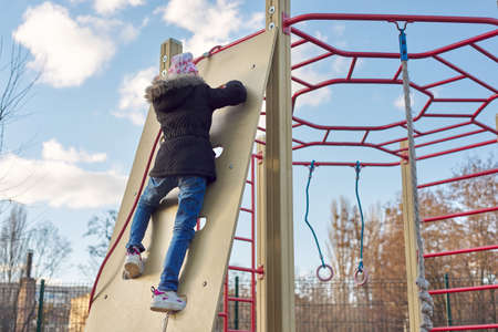 Girl child of 8, 9 years old on outdoor sports equipment, childrens playground. Spring season, girl in jacket and hatの写真素材