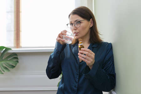 Nervous disorder, depression, nervousness, age-related health problems. Mature woman dripping drops with glass of water, drinking sedative medicine, at home in pajamas, copy spaceの写真素材