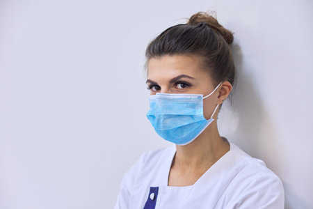 Young serious woman doctor in medical mask looking at camera, background wall to the hospital, copy...