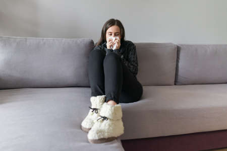 Sad sick young woman with handkerchief. Female with cough, snot, fever sitting at home on sofa, copy spaceの写真素材