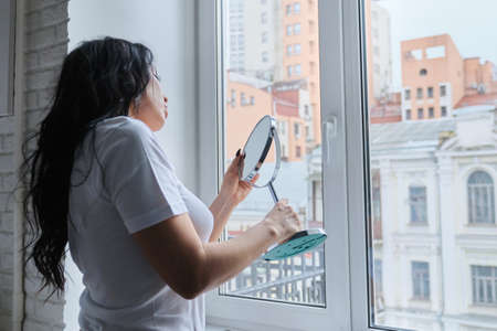 Beautiful brunette woman with long wavy hair admires herself in mirror, female at home near window looks at makeup mirror, copy spaceの写真素材