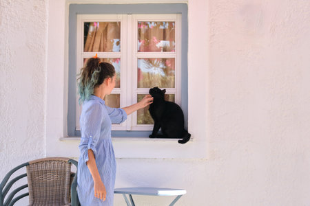 Teenager girl touching beautiful black cat sitting on the window sill outdoor, sunny summer day,...