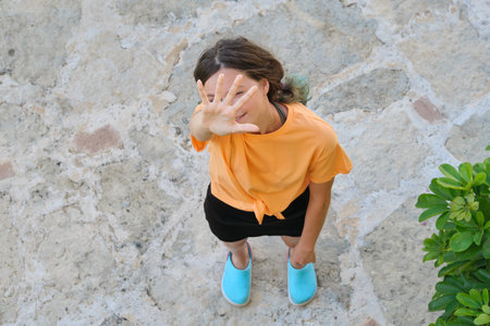 Teenager girl waving, welcomes, looks up at camera, shows hello gesture. Stands on sidewalk on...