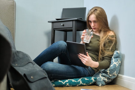 Young woman sitting at home on floor with digital tablet and drinking glass of water. Girl studies,...