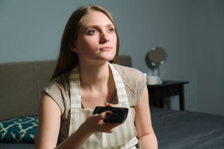 Young beautiful woman with remote control, serious female sitting at home and watching tv.