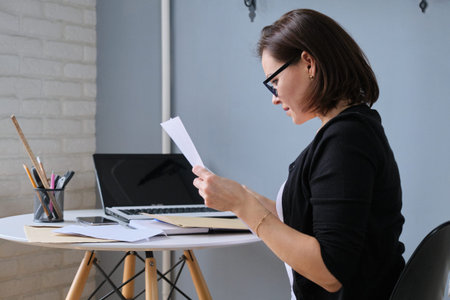 Mature business woman sitting at desk in office reading paper document, business correspondence,...