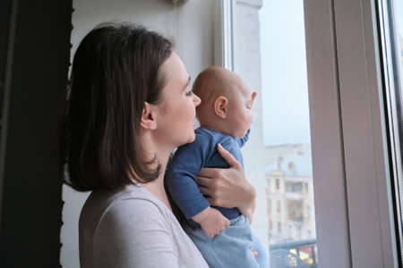 Young woman with little baby son in her arms. Mother and baby stand at home near window, look out the windowの写真素材