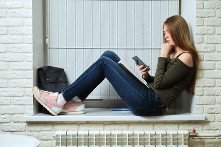 Girl student with smartphone, young beautiful woman with backpack notebooks sitting on windowsill...