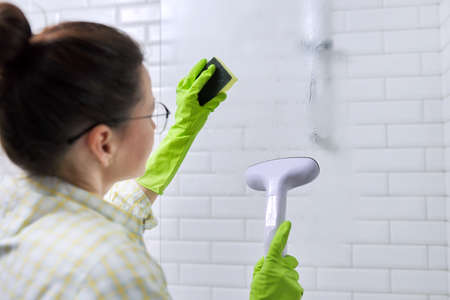 Woman housewife doing house cleaning in bathroom, female washing glass with shower steam. Using steam cleaner for quick cleaningの写真素材