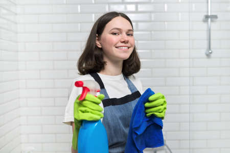 Portrait of girl doing cleaning in bathroom. Teenager in apron gloves with detergent and rag sponge smiles looking at camera. Housekeeping, cleanliness at home, service, young people conceptの写真素材