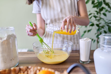 Close-up process of preparing orange pancakes, female hands pouring orange juice into a bowl with...