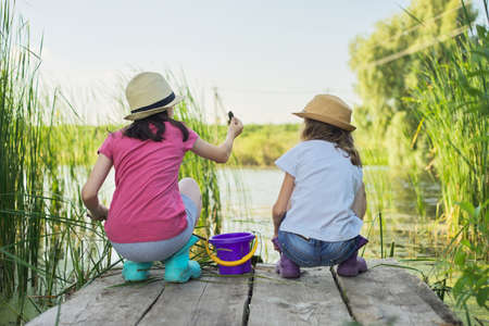 Children two girls together playing with water on lake on wooden pier in reeds. Kids with bucket picking sticks of algae and water snails. Nature, leisure, friendship, summer vacation conceptの写真素材