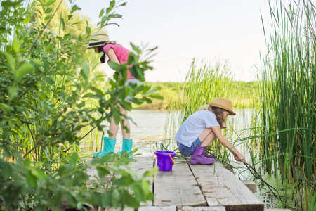 Children two girls together playing with water on lake on wooden pier in reeds. Kids with bucket picking sticks of algae and water snails. Nature, leisure, friendship, summer vacation conceptの写真素材