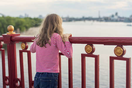 Child blonde with long hair standing back on pedestrian bridge over river. Girl dreamily watching and enjoying sunset on water surfaceの写真素材