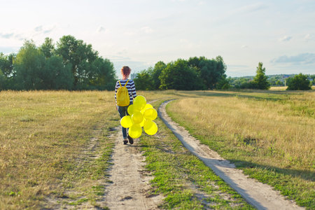 Teenager girl with backpack and yellow balloons running on country road, rear view. Happy girl walking forward, sky in the clouds, meadow, nature backgroundの写真素材